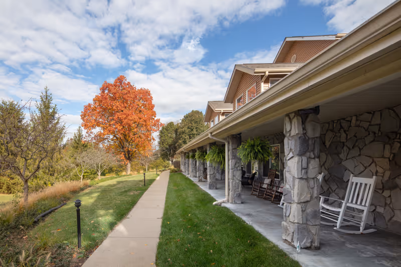 A stone building with a covered porch featuring several rocking chairs and hanging ferns. A concrete walkway runs alongside the porch, bordered by green grass and trees, including one with bright orange autumn leaves under a partly cloudy sky.