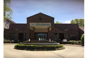 Front exterior view of The Altenheim Senior Health Care Community building with a circular driveway and landscaped bushes in front under a clear blue sky.