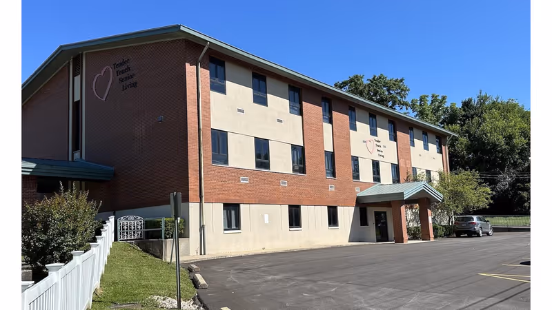 Front exterior of a three-story brick-and-stucco building labeled "Tender Touch Senior Living" with a parking lot and entrance canopy.