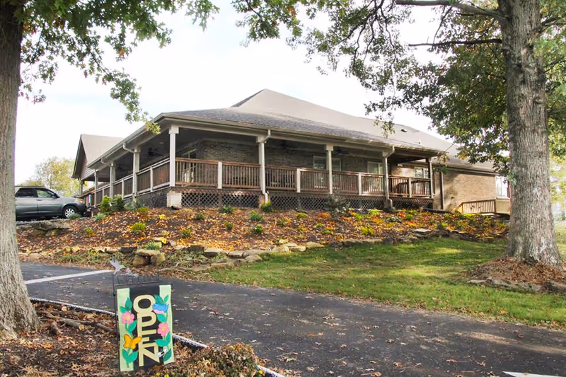 Exterior view of a single-story brick building with a large covered porch and wooden railing, surrounded by trees and a landscaped garden with flowers. A paved driveway leads up to the building, and a colorful sign with the word 'OPEN' is placed near the driveway entrance.