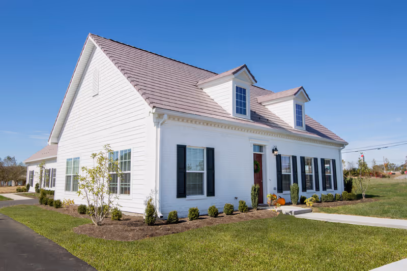 Front view of a white single-story house-like building with dormer windows, a red door, black shutters, and landscaped lawn under a clear blue sky.