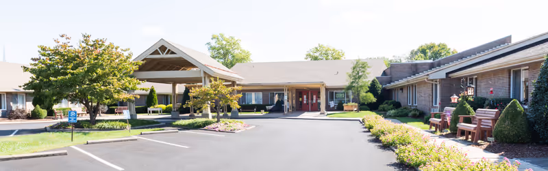 Front exterior view of Christian Health Center Hopkinsville showing a single-story building with a covered entrance, parking spaces, landscaped greenery, and benches along the walkway.