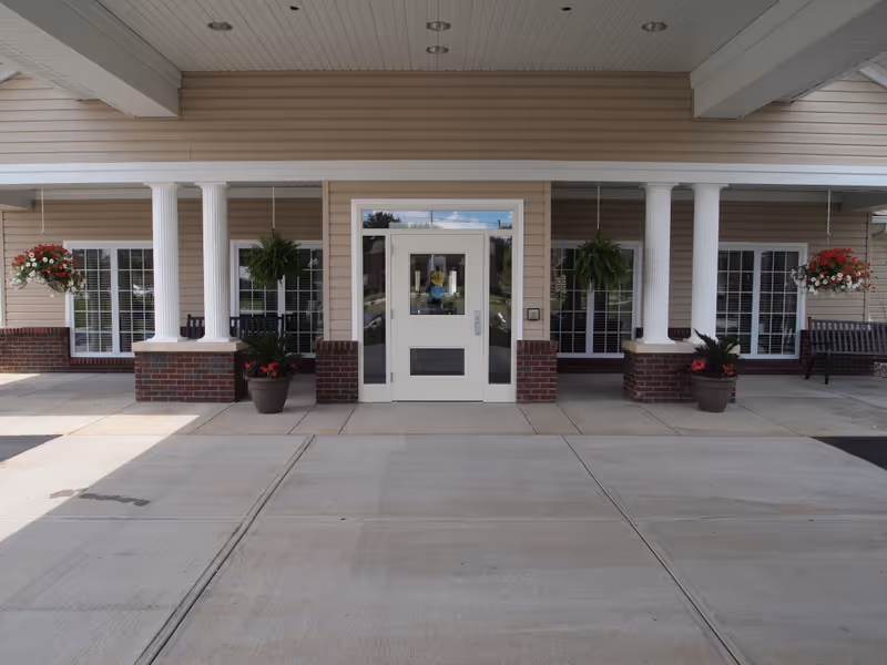 Front entrance of Westport Place Health Campus featuring a covered drop-off area with white columns, hanging flower baskets, potted plants, and benches on either side of the door.