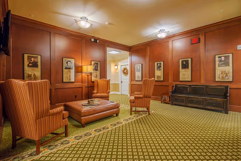 A cozy sitting area with three striped armchairs arranged around a large brown leather ottoman. The walls are paneled with dark wood and decorated with framed portraits. The floor is covered with a green patterned carpet. There is a lamp between two chairs and a black wooden bench against the wall. A doorway with a wreath on the door is visible in the background.