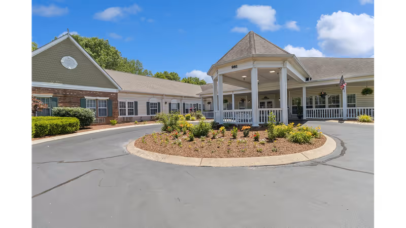 Front entrance of a single-story senior living building with a covered portico, circular driveway, and landscaped center island.