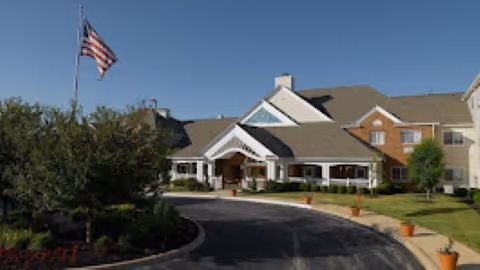 Front exterior view of a senior living facility building with a driveway, landscaped greenery, and an American flag on a flagpole.