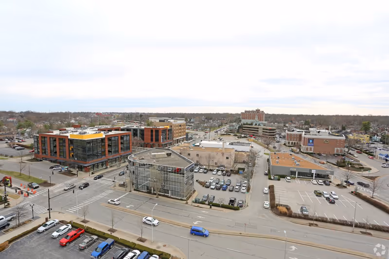 Aerial view of a commercial area with multiple buildings, parking lots, and streets. The scene includes a modern multi-story building with large windows, several smaller buildings, and numerous parked cars. The sky is overcast.