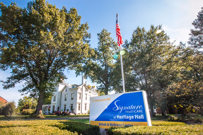 White two-story building with a large blue 'Signature HealthCARE at Heritage Hall' sign and flagpole.