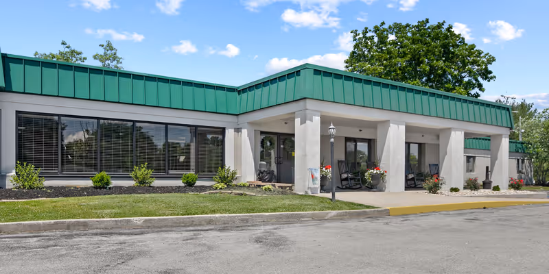 Exterior view of a single-story building with a green roof and large windows. The entrance features a covered porch with rocking chairs and potted plants. There is a small lawn and a paved driveway in front of the building under a blue sky with some clouds.