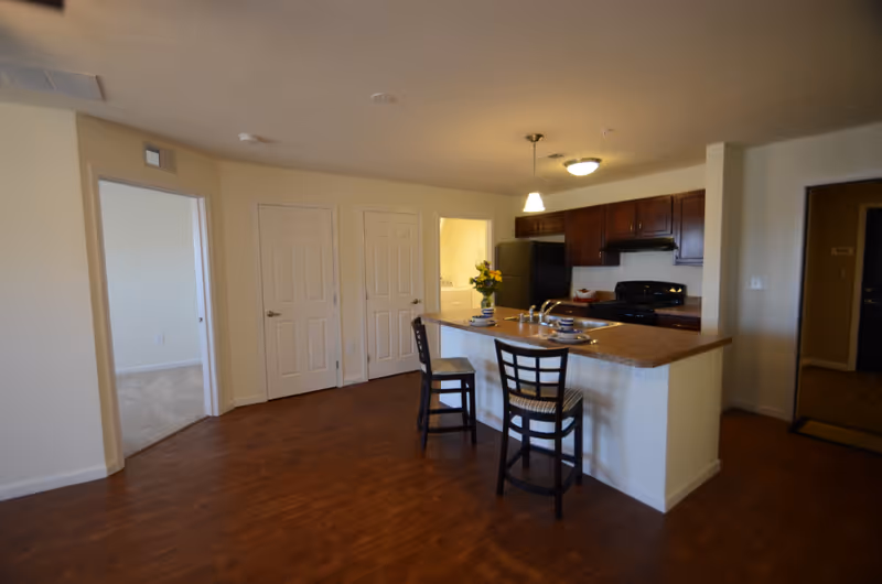 Interior view of a senior living apartment kitchen area with a countertop island featuring two chairs, a sink, and place settings. The kitchen has dark wood cabinets, a black refrigerator, and a stove. There are two closed doors and an open doorway leading to another room with carpeted flooring.