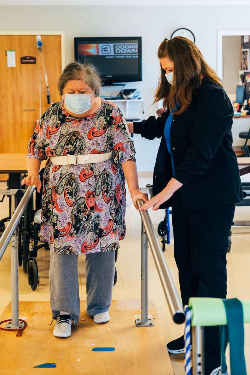 An elderly woman wearing a face mask and a patterned dress is walking with the support of parallel bars in a rehabilitation or therapy room. A caregiver or therapist, also wearing a face mask, is assisting her by holding her arm. In the background, there is a television mounted on the wall and a wheelchair.