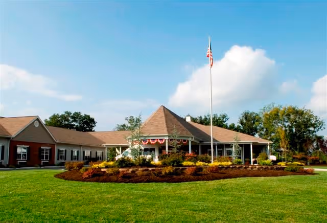 Exterior view of a single-story senior living facility building with a peaked roof, surrounded by a well-maintained lawn and landscaped flower beds. An American flag is flying on a flagpole in front of the building under a partly cloudy sky.