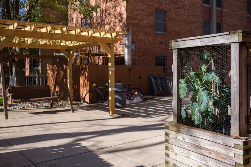 Outdoor patio area with a wooden pergola and a wooden swing bench underneath. There are two bicycles parked near a brown fence, and a wooden planter box with a plant protected by chicken wire is in the foreground. A brick building is visible in the background.