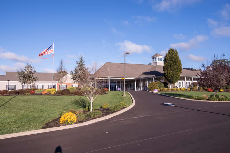 Exterior view of Morning Pointe of Russell senior living facility with a paved driveway, landscaped lawn with bushes and trees, an American flag on a flagpole, and a clear blue sky.