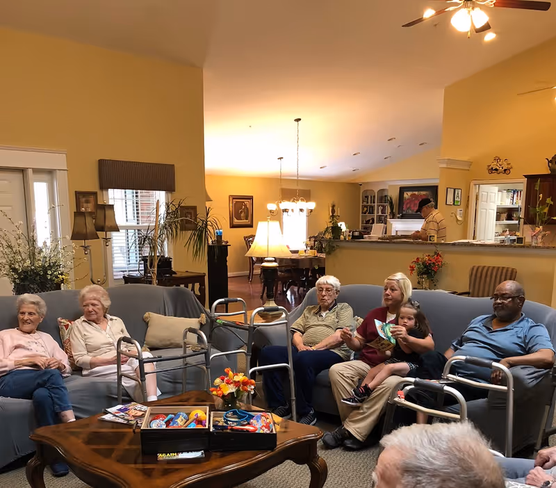 A bright communal living room in a senior home with several elderly residents and a caregiver holding a child seated on sofas surrounded by walkers and lamps.