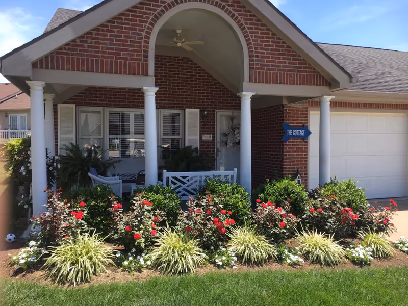Front exterior of a brick cottage-style residence with a covered porch, white columns, and a landscaped flowerbed with red roses.