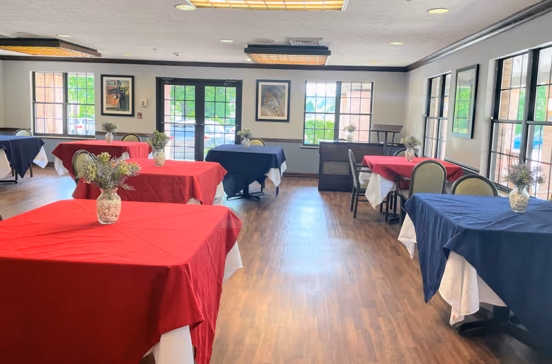 A bright dining room with several tables covered in red and blue tablecloths, each with a small vase of flowers. The room has large windows letting in natural light, wooden flooring, and framed pictures on the walls.