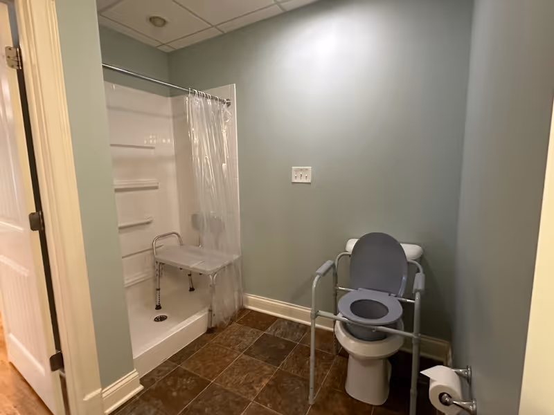 A bathroom with a walk-in shower that has a clear plastic curtain and a white shower chair inside. There is a toilet with a raised seat and arm supports for accessibility. The walls are painted light green and the floor is covered with brown tiles.