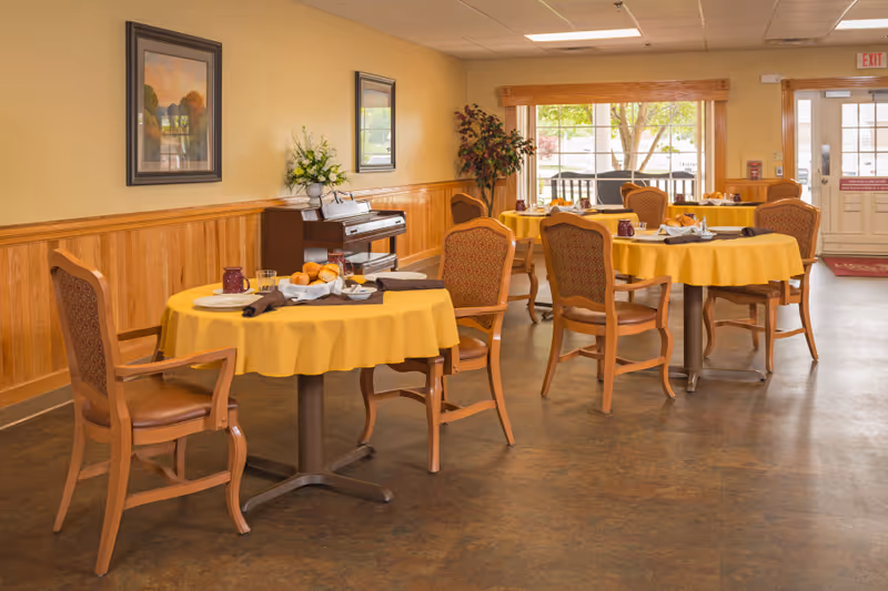 A dining room with round tables covered in yellow tablecloths, each set with plates, glasses, and napkins. Wooden chairs with cushioned seats surround the tables. A piano with a flower arrangement on top is against the wall, which is decorated with framed paintings. Large windows and a door let in natural light, and a potted plant is in the corner.