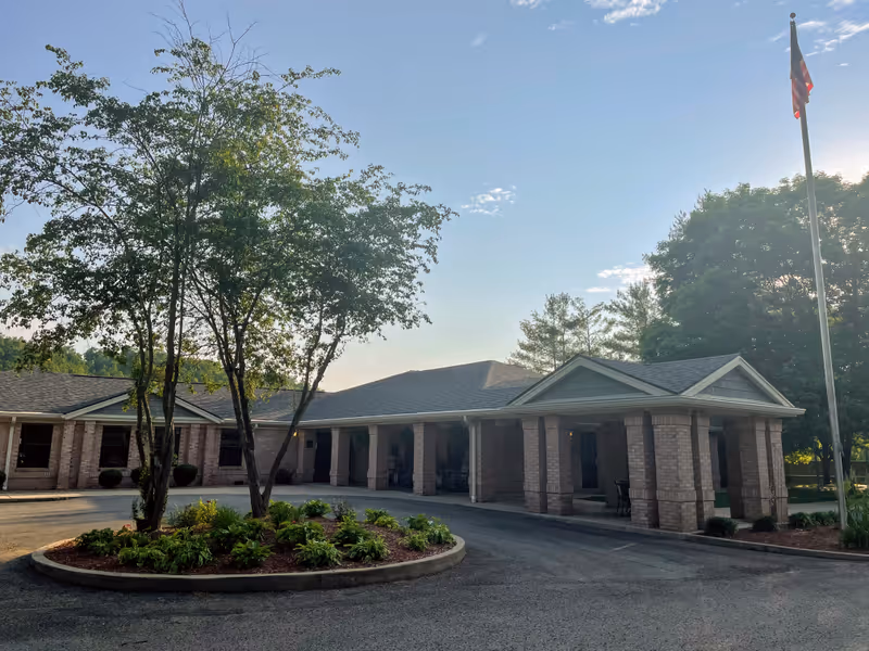 Exterior view of Elliott Nursing and Rehabilitation facility showing a single-story brick building with a covered entrance supported by columns. There is a circular landscaped area with trees and plants in front, and an American flag on a tall flagpole to the right. The sky is clear with some clouds and the sun is low, casting shadows.