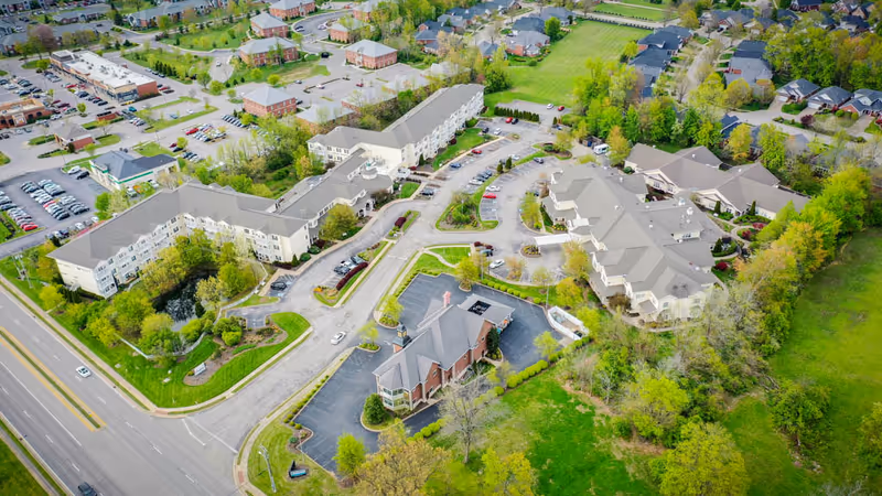 Aerial view of Arcadia Senior Living Louisville facility showing multiple large buildings surrounded by parking lots, roads, green lawns, and trees. The complex is located near a main road and residential neighborhood.