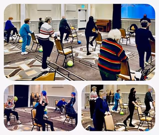 A group of elderly individuals participating in a seated and standing exercise class in a spacious room with patterned carpet and white walls. They use chairs for support and colorful exercise balls as part of their routine.