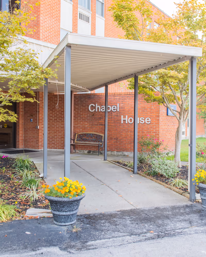 Entrance to Chapel House Corbin, a Christian Care Community, showing a covered walkway supported by metal poles, a brick wall with the words 'Chapel House', a bench, and surrounding greenery with yellow flowers in planters.