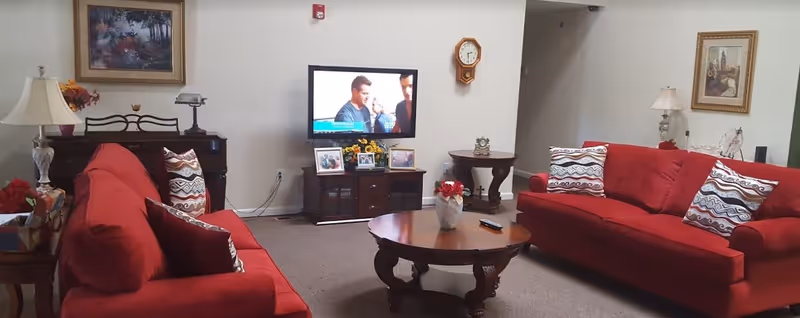 A cozy living room with two red sofas facing each other, each adorned with patterned throw pillows. A round wooden coffee table with a vase of flowers and a TV remote sits in the center. A flat-screen TV is mounted on the wall above a wooden cabinet displaying framed photos and a small flower arrangement. The room also features a wooden side table with a small clock, a wall clock, two table lamps, and framed artwork on the walls.