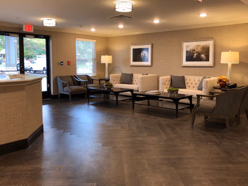 A cozy senior living facility lounge area with two beige tufted sofas, two gray armchairs, two black coffee tables with decorative items, two floor lamps, framed horse artwork on the wall, and a reception desk near a glass door with an exit sign.