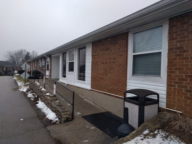 Brick senior living facility exterior with ramp, windows, trash can, and patched snow.