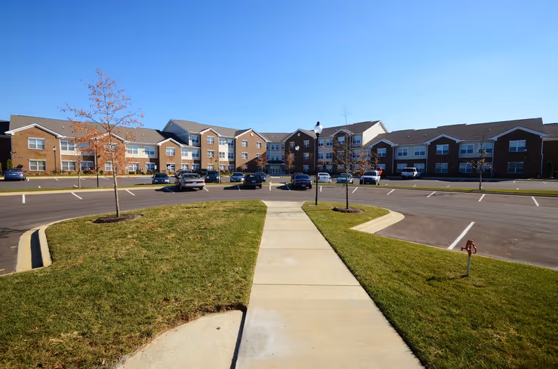Wide view of the exterior of Hamburg Senior Residence, showing a large three-story brick and siding building with multiple windows. In front of the building is a paved parking lot with several parked cars, a sidewalk leading towards the entrance, and small trees planted in grassy areas.