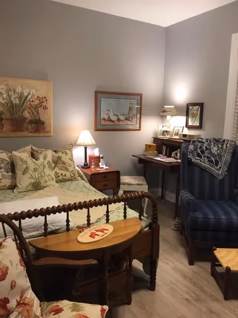 Cozy bedroom with a wooden spindle bed, floral bedding, bedside tables with lamps, a blue upholstered armchair, and a small desk.