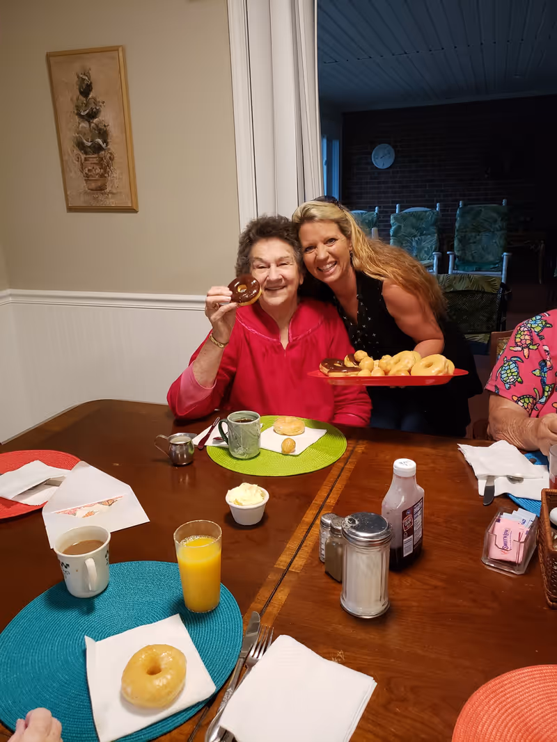 An elderly woman in a red top sitting at a dining table holding a chocolate glazed donut and smiling. A younger woman with long blonde hair is standing behind her, smiling and holding a red tray filled with various donuts. The table has colorful placemats, cups of coffee, a glass of orange juice, condiments, and napkins. Another person is partially visible on the right side of the image. The setting appears to be a cozy dining area in a senior living facility.