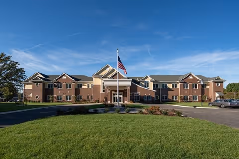 Front exterior view of a two-story brick assisted living facility with a central entrance, an American flag on a flagpole in front, a well-maintained lawn, and a clear blue sky.