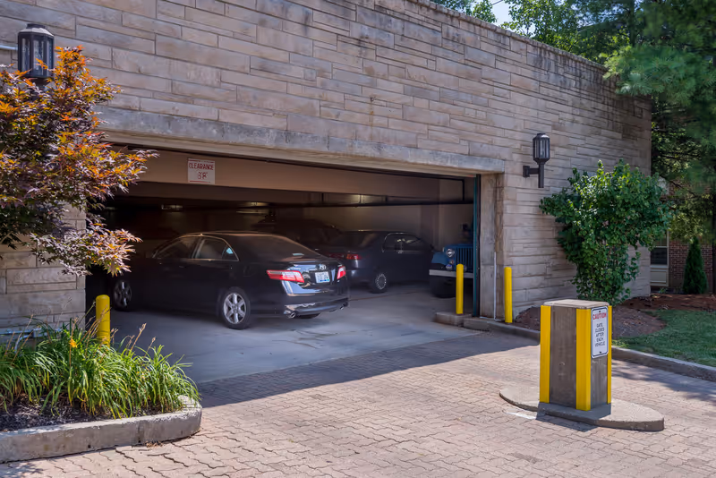 Open stone-faced parking garage entrance with cars parked inside, plants and yellow bollards in front.