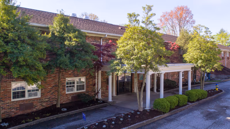 Exterior view of a brick building with a covered entrance supported by white columns. There are several trees and bushes along the walkway leading to the entrance. The sky is clear and the scene is well-lit by daylight.