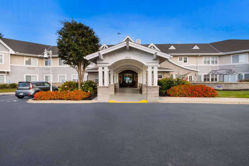 Front entrance of a two-story senior living building with a covered porte-cochere, landscaped flowerbeds, and parked cars.