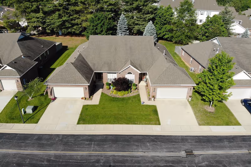Aerial view of a single-story residential building with a gray shingled roof and brick exterior. The building has two attached garages, a well-maintained green lawn, and landscaped bushes and trees in front. It is situated in a neighborhood with similar houses and surrounded by tall trees in the background.