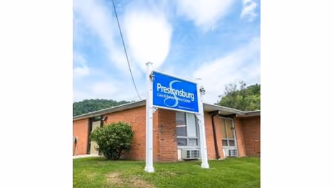 Exterior view of a single-story brick building with a blue sign in front that reads 'Prestonsburg Care & Rehabilitation Center' under a partly cloudy sky.