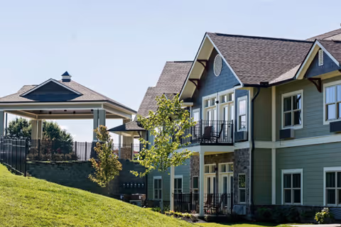 Exterior view of a senior living facility building with green siding, multiple windows, and balconies. There is a grassy hill in the foreground and a covered pavilion structure to the left. Trees and a clear blue sky are visible in the background.