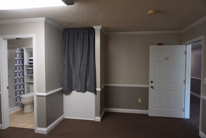 Interior view of a room showing a white entrance door numbered 17, an open bathroom with shower and toilet, and a gray curtained window.