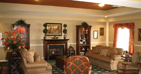 A cozy living room with two beige sofas, a wooden coffee table, a fireplace with a framed painting above it, two tall wooden bookshelves with decorative items, and a window with red curtains letting in natural light.