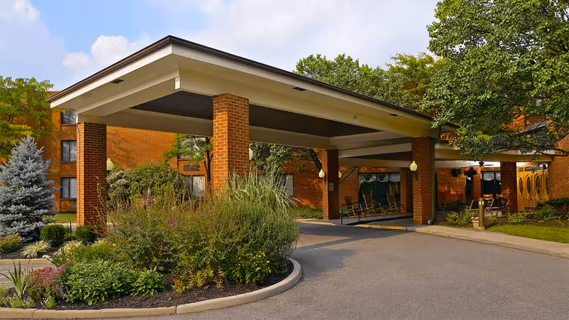 Entrance area of Celebration Villa of Highland Crossing featuring a covered drop-off with brick pillars, surrounded by landscaped greenery and trees, with a clear sky above.