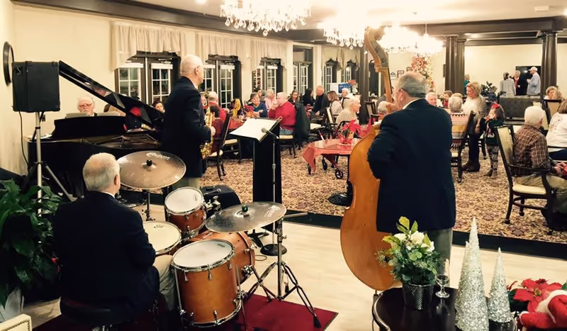 A live band with a drummer, saxophonist, and double bass player performing in a room filled with elderly people seated at tables, enjoying the music. The room is decorated with chandeliers, plants, and festive holiday decorations.