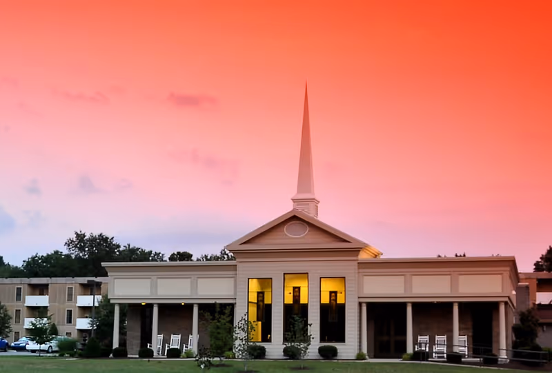 Exterior view of a building with a tall white steeple against a vibrant pink and orange sunset sky. The building has large windows with warm yellow light shining through and a porch area with white rocking chairs. There are trees and shrubs in front of the building and an apartment complex visible in the background.