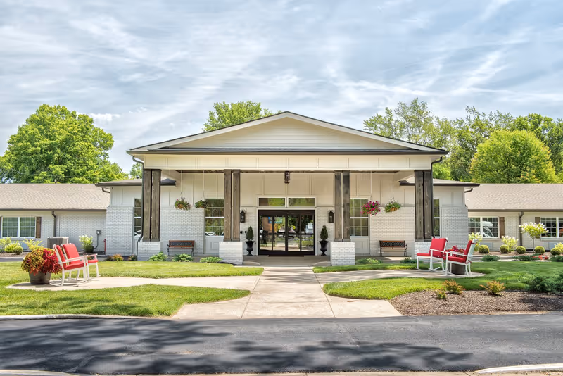 Front exterior view of a single-story senior living facility building with a covered entrance supported by wooden pillars. There are benches and red cushioned chairs on either side of the walkway leading to the entrance. The building is surrounded by well-maintained green lawns, plants, and trees under a partly cloudy sky.
