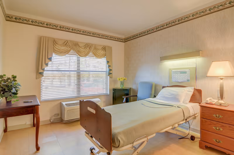 A well-lit patient room with a single hospital bed covered in beige bedding, a wooden nightstand with a lamp and telephone, a blue armchair, a small table with a plant, and a window with beige curtains and blinds.