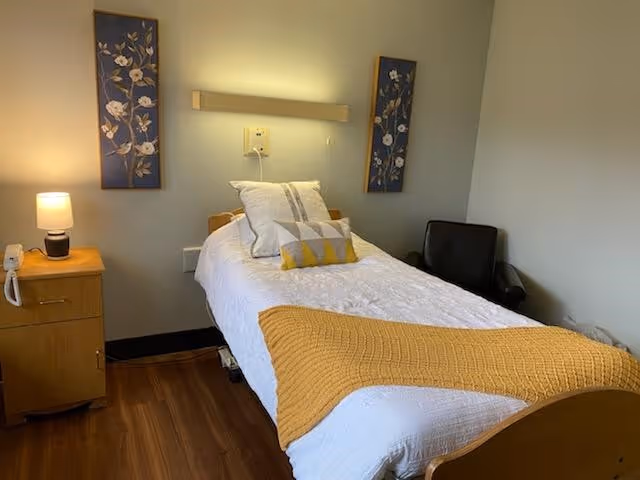 A single bed with white bedding and a mustard yellow throw blanket is positioned against a light gray wall. Two floral paintings hang above the bed. To the left of the bed is a wooden nightstand with a lamp and a telephone. A black armchair is placed in the corner to the right of the bed. The floor is wooden.