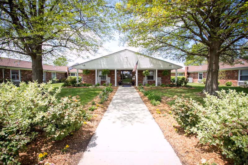 Front exterior view of Bluegrass Care & Rehabilitation building with a concrete walkway leading to the entrance. The building is single-story with brick walls and a white gabled roof. There are two large trees and landscaped bushes and flowers on either side of the walkway. The entrance has a covered porch with rocking chairs and hanging plants, and an American flag is displayed near the door.