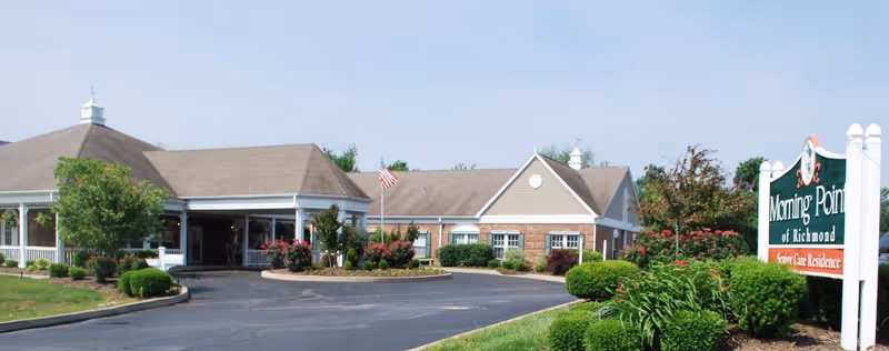 Exterior view of Morning Pointe of Richmond senior care residence showing a single-story building with a covered entrance, surrounded by well-maintained landscaping including bushes, flowers, and trees, with an American flag near the entrance and a clear sky above.